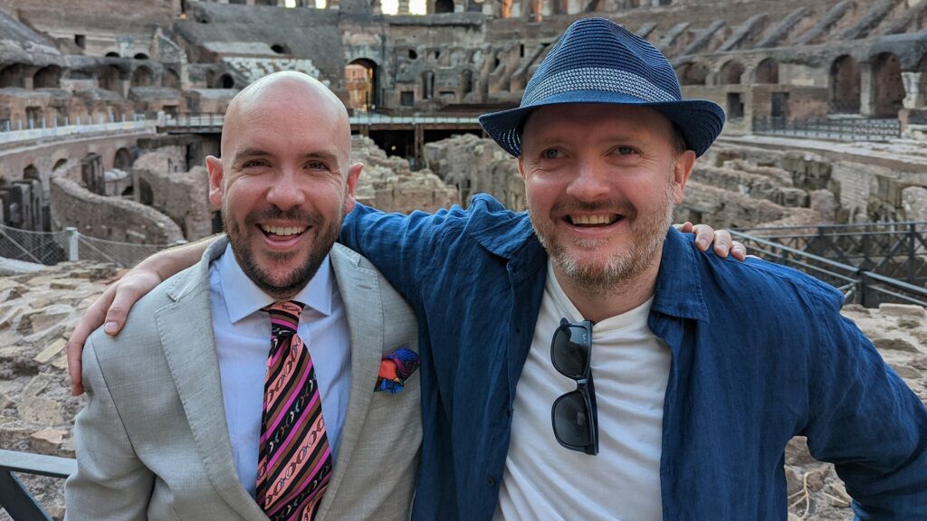 a bald white man with a blue collared shirt and red stripped tie stands next to a white man who is wearing a blue hat, blue jacket with a white t-shirt.