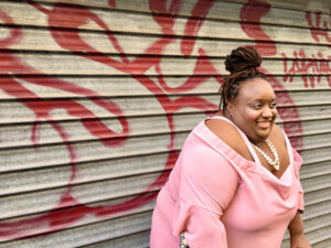 A Black woman with Black hair pulled up into a bun wearing a pink blouse stands in front of a silver garage door with graffiti.