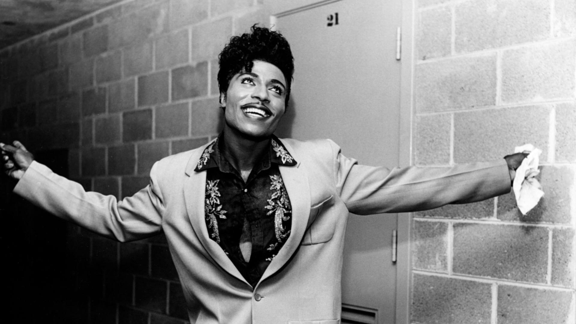 A photo Black and shite of Little Richard - a Black man with short Black hair and a mustache wearing a light suit and leaf patterned button down shirt.