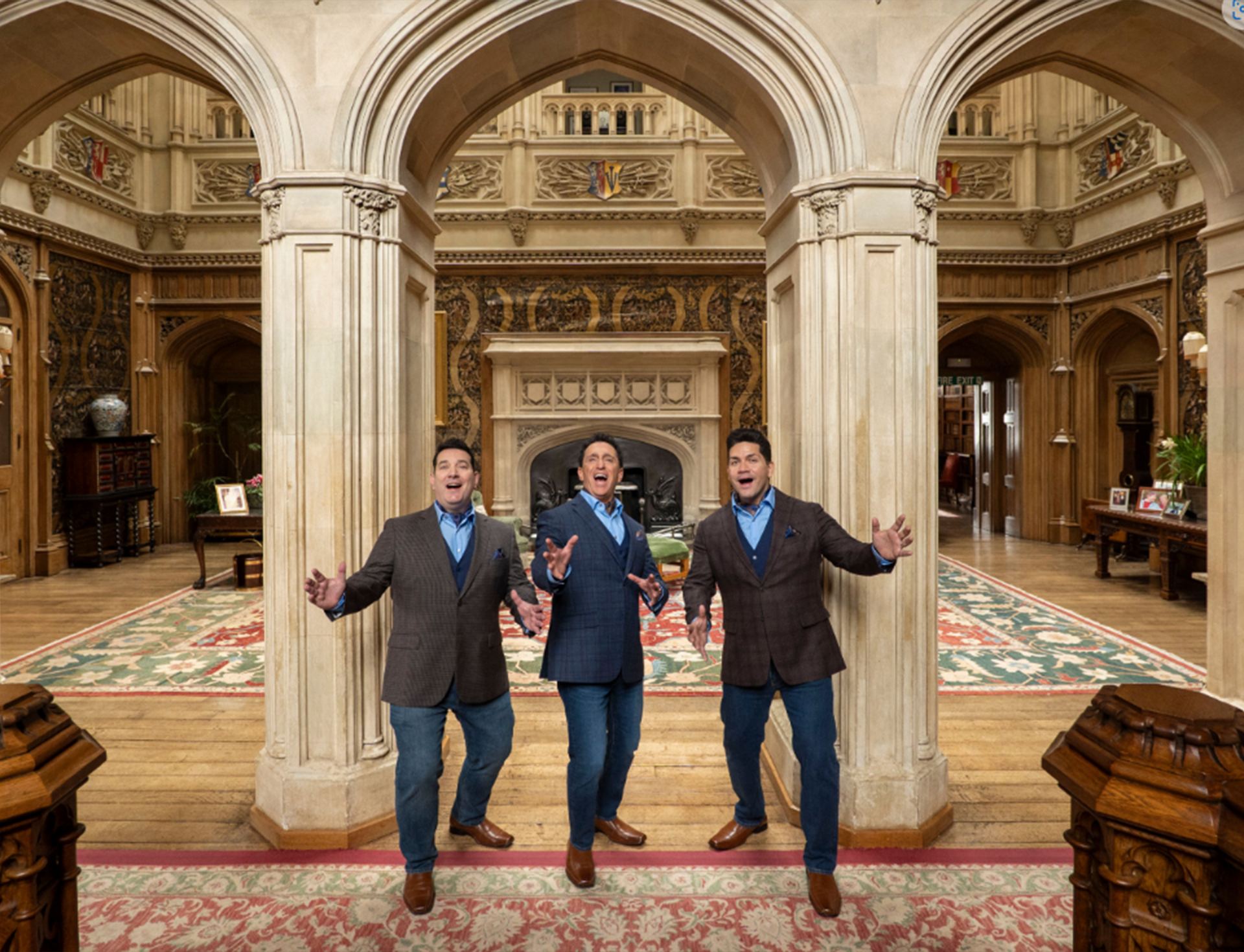Three white men in dark blazers and jeans stand in front of a great room in a castle with their arms outstretched