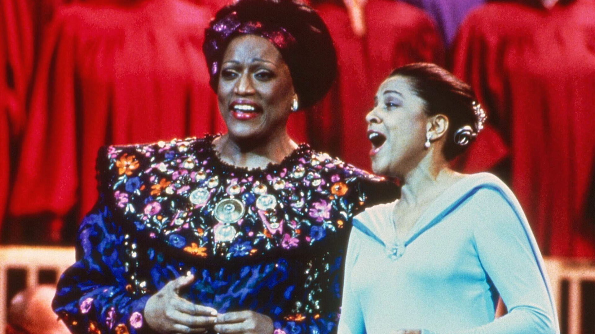 Two Black woman stand on stage singing. The woman on the left wears a blue dress with a floral collar and the woman on the right ears a light blue dress.