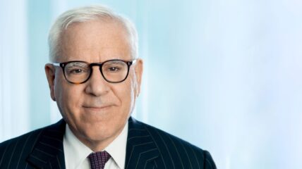 A photo of David Rubenstein - a white male with short gray hair wearing black rimmed glasses and a suit and tie.