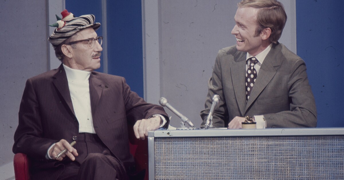 Two white men sit at a desk talking to each other as part of a TV show.