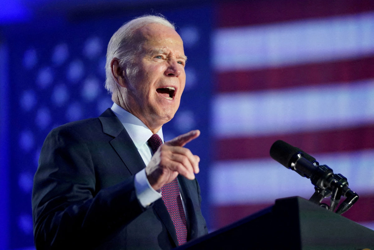 President Biden - an older white male with short gray hair wearing a blue blazer, white collared shirt and red and black dotted tie stands at a podium making a speech. In the background is the American flag.