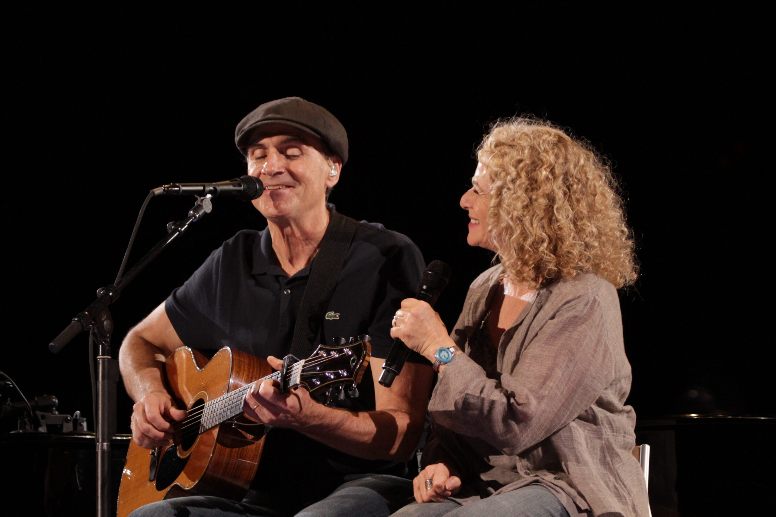 A white man wearing a Black t-shirt and Black cap plays the guitar in front of a mic. A white woman with wavy gray shoulder length hair sits next to him with a microphone in hand.