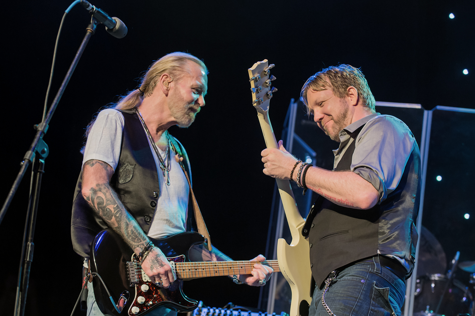 A white man with blonde hair pulled back in a pony tail and a blond beard wearing a gray tee and leather vest plays an electric guitar alongside another white man with short brown hair wearing a gray collared shirt and Black vest also playing an electric guitar.