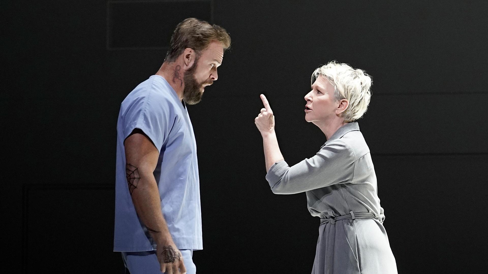 A white man with brown short hair, beard and mustache wearing prison scubs stands before a white woman with short blonde hair wearing a gray dress. She is pointed her finger at him.