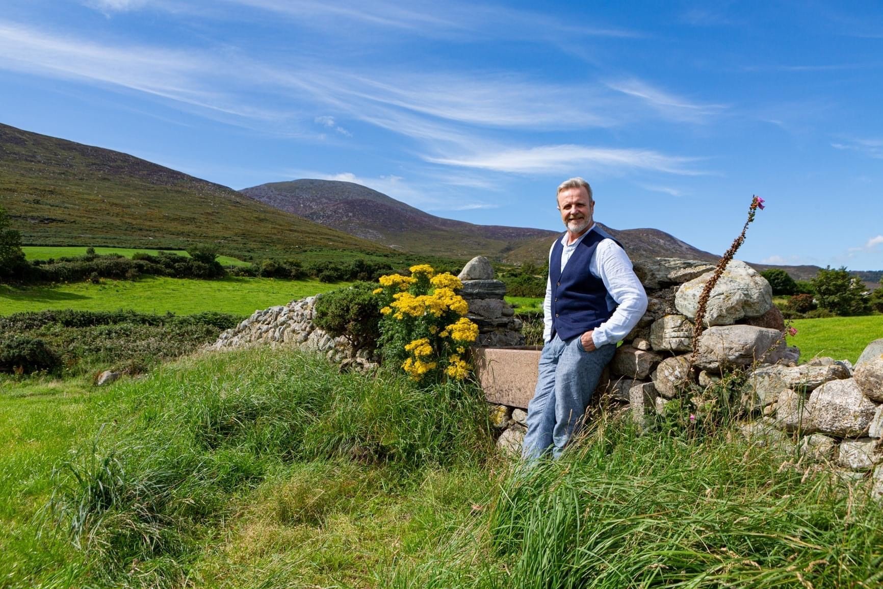 A white man with gray hair and facial hear wears a blue vest, light blue collared shirt and jeans stand in front of a grassy fiend with flowers and mountains in the background