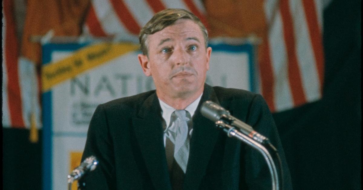 A white man with short brown hair wearing a navy suit, white shirt and blue tie sits behind two microphones