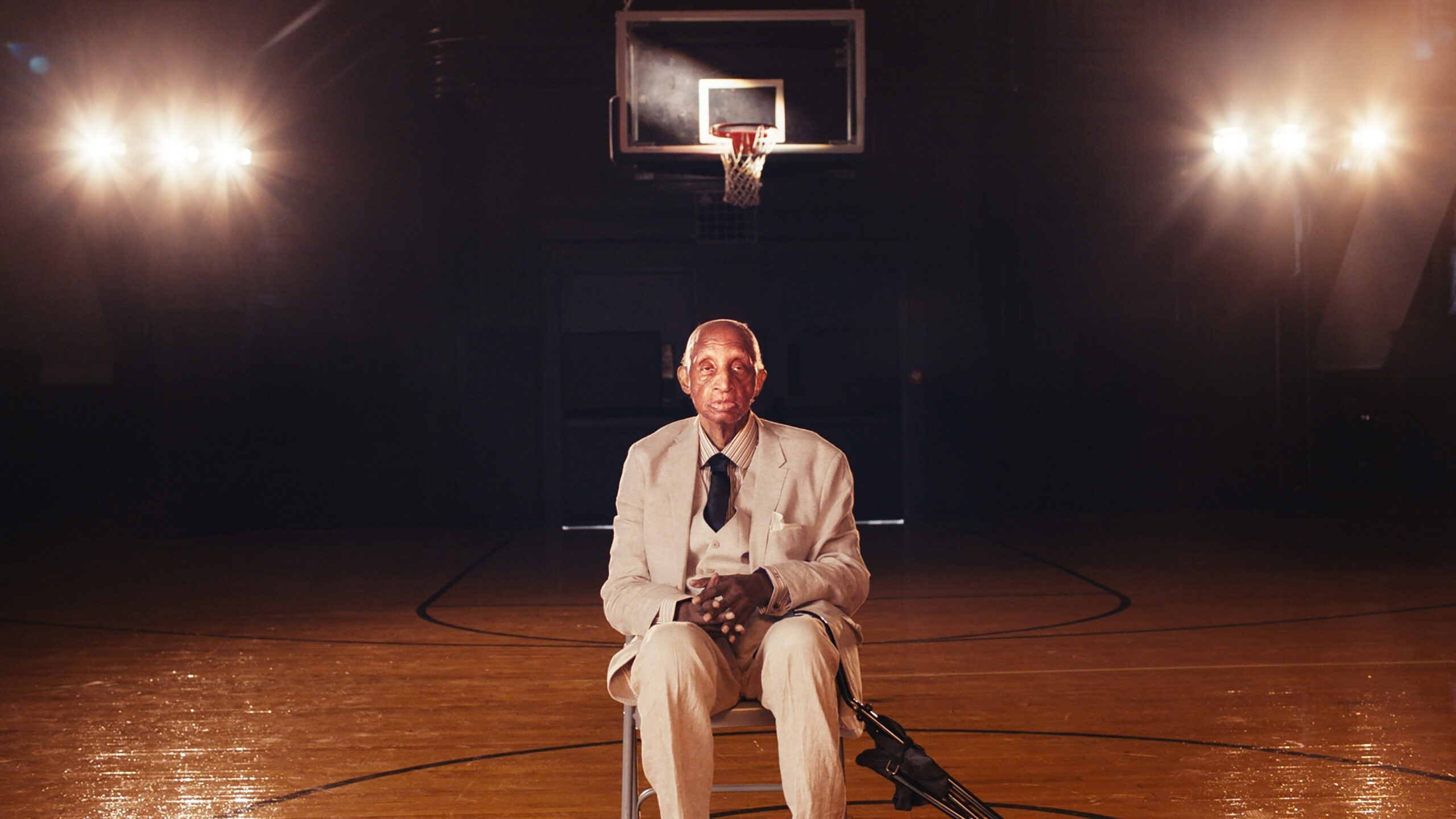 An older Black man in a tan suit and brown tied sits on chair on a empty basketball court