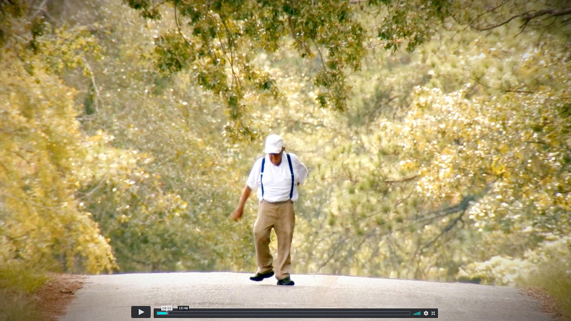 An white man in a white baseball cap, white tee, khaki pants and blue suspenders walks along street lined with trees