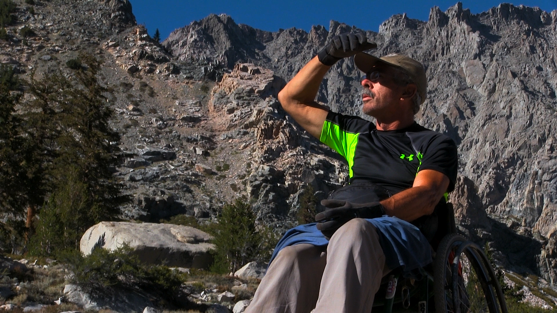 A man in a wheel chair with a baseball cap and an athletic Blacka nd green shirt in a wheel chair. Mountains are behind him