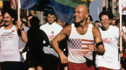 A white male in a tank top with the American flag and red running shorts running with friends behind him