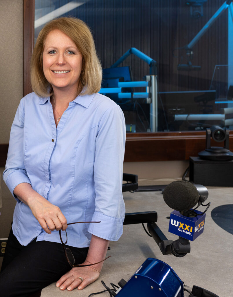 A white woman with blonde shoulder length hair wearing a blue button down shirt sits in a radio studio.
