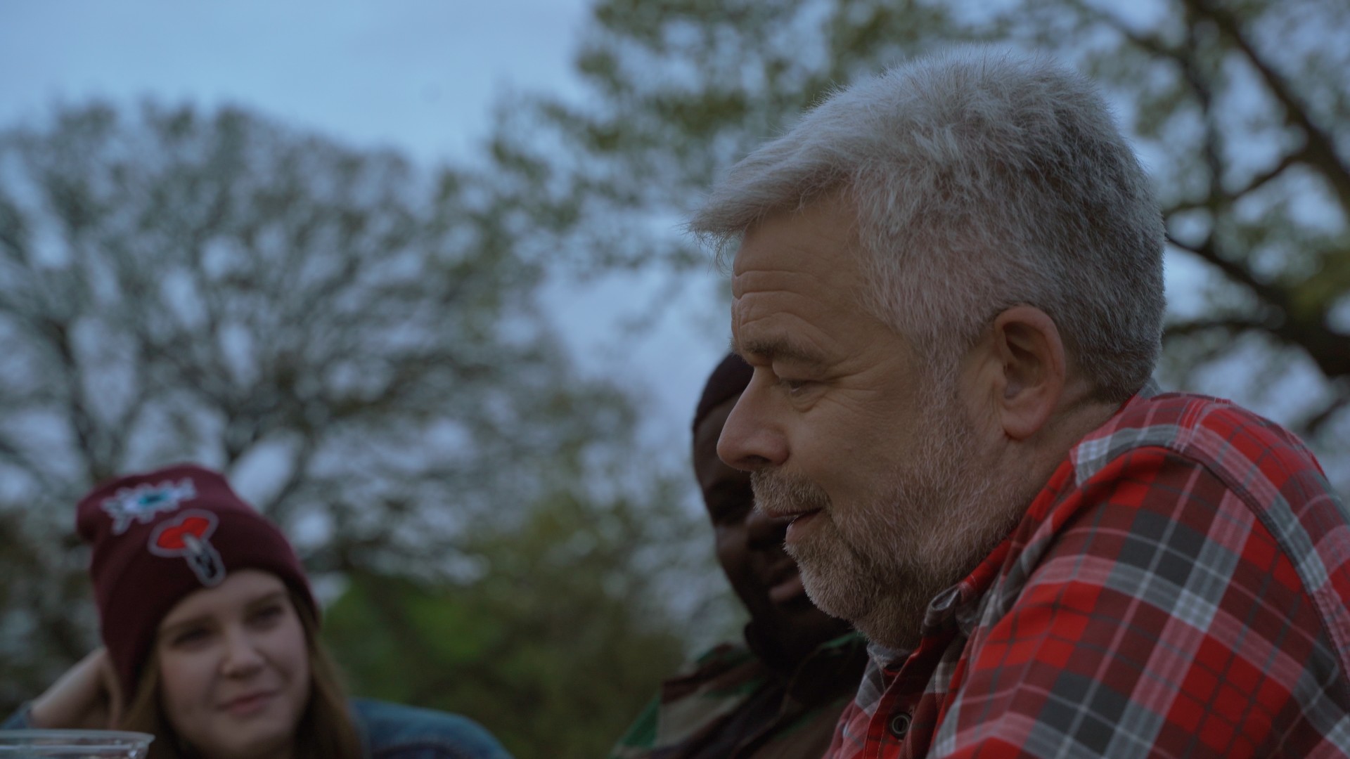 A males with short gray hair wearing a red and Black flannel sit next to a young white woman and young Black man talking