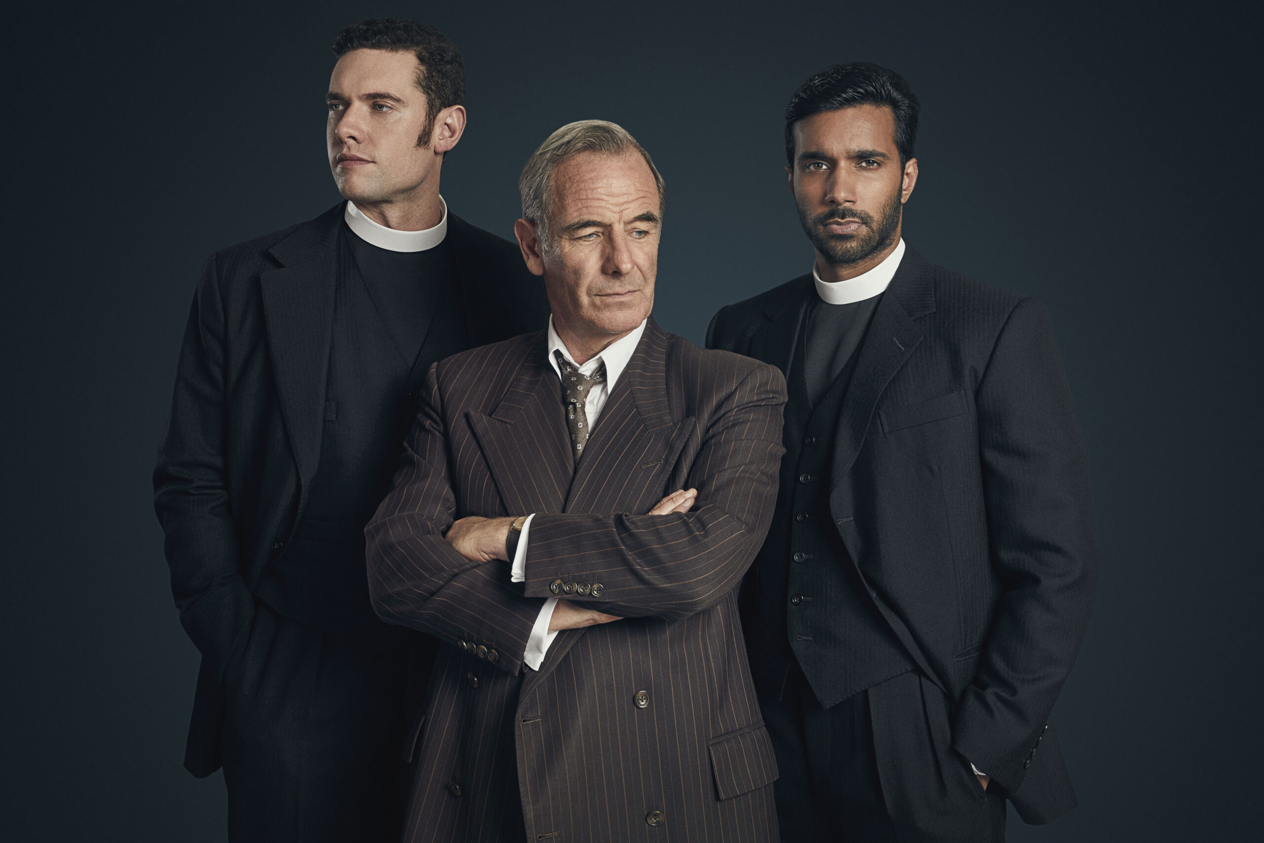 Three men stand together - the middle man wearing a dark suit, tie and white collared shirt. The men on each side of him are wearing vicar collars.