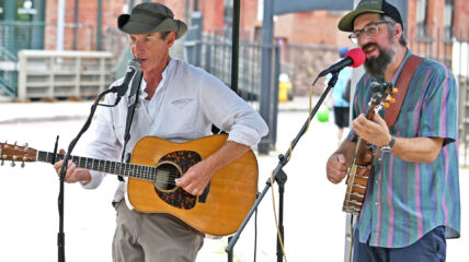 Two white men singing. The man on the left is strumming an acoustic guitar.