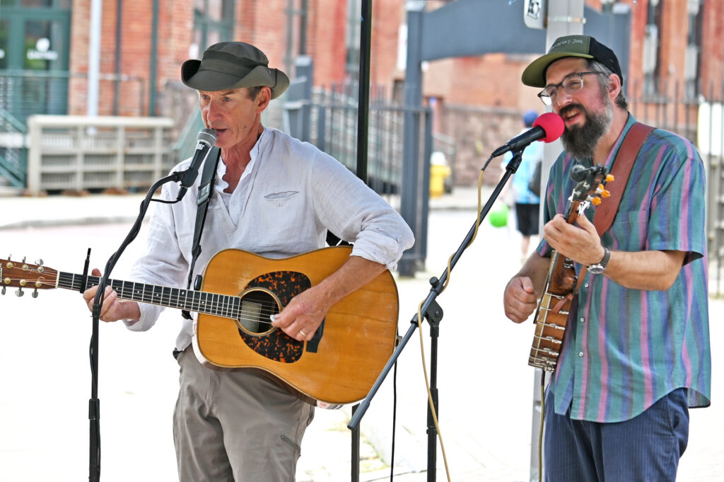 two white men singing into microphones. The man on the left is strumming an acoustic guitar