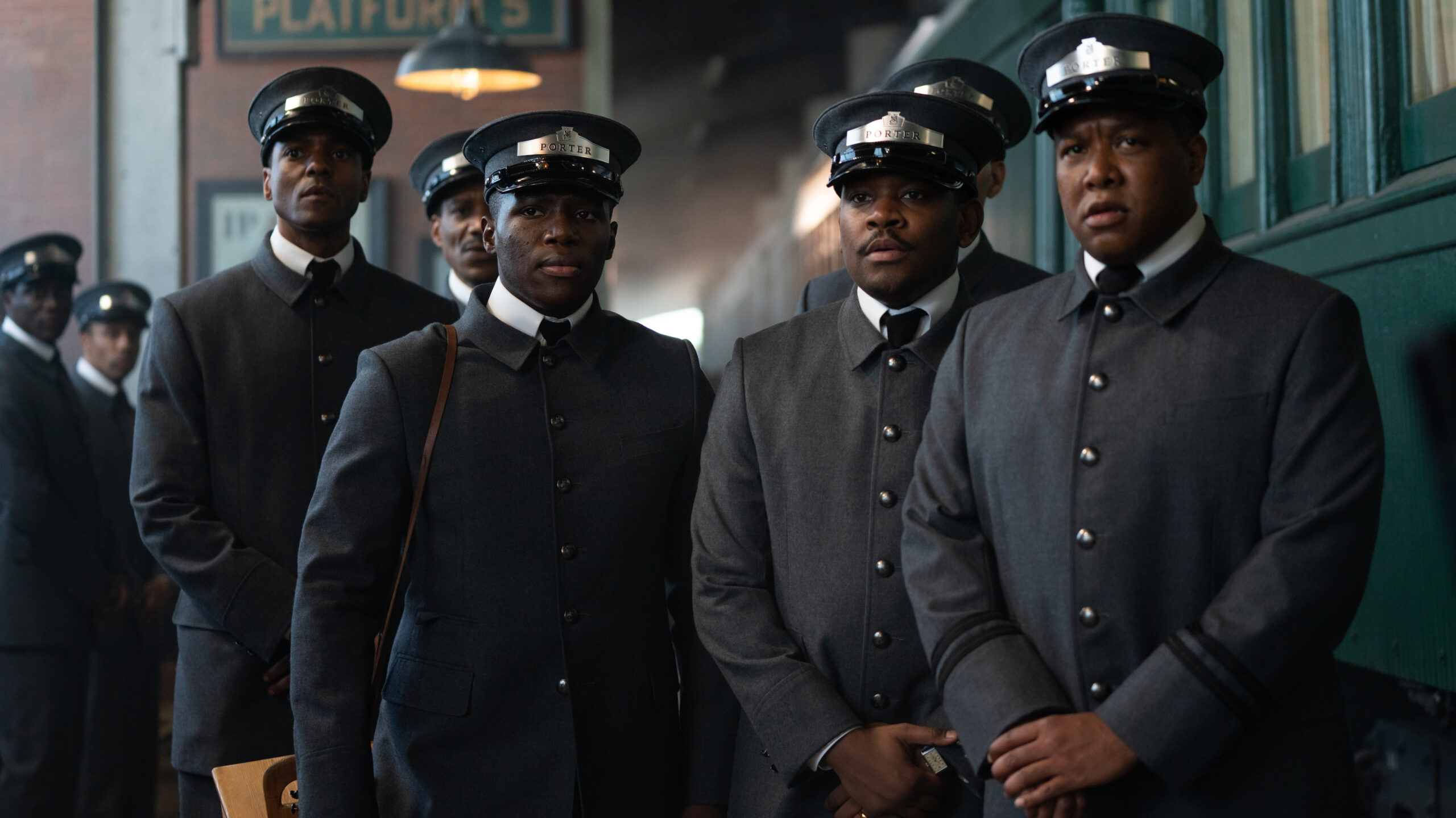 Two Black men in Porter uniforms stand in front of a train. Additional porters in the background.