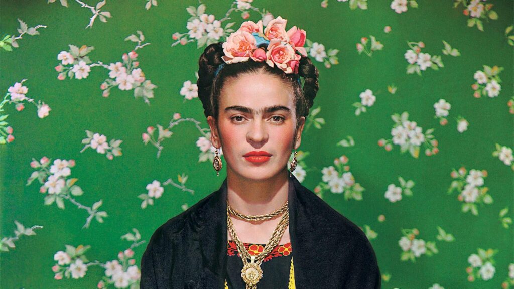 A painting of artist Frida Kahlo - She has her Black hair up in a bun with a big flower tucked into the front of her hair. She is wearing a floral blouse with a Black sweater.