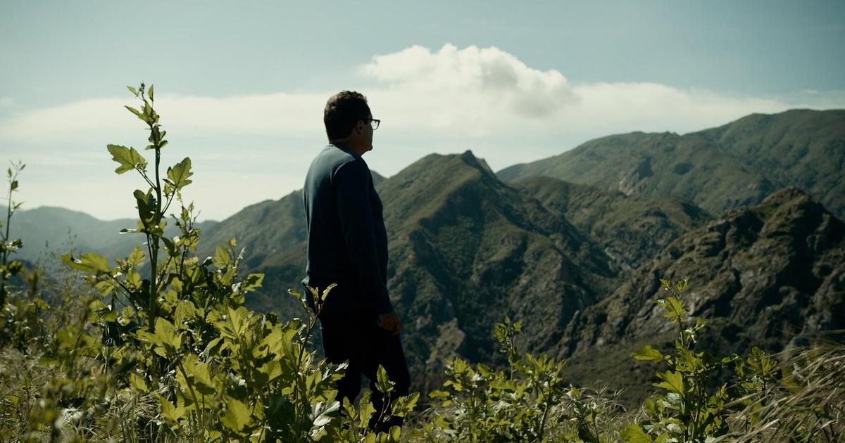A white male stands in a field with mountains in the background.