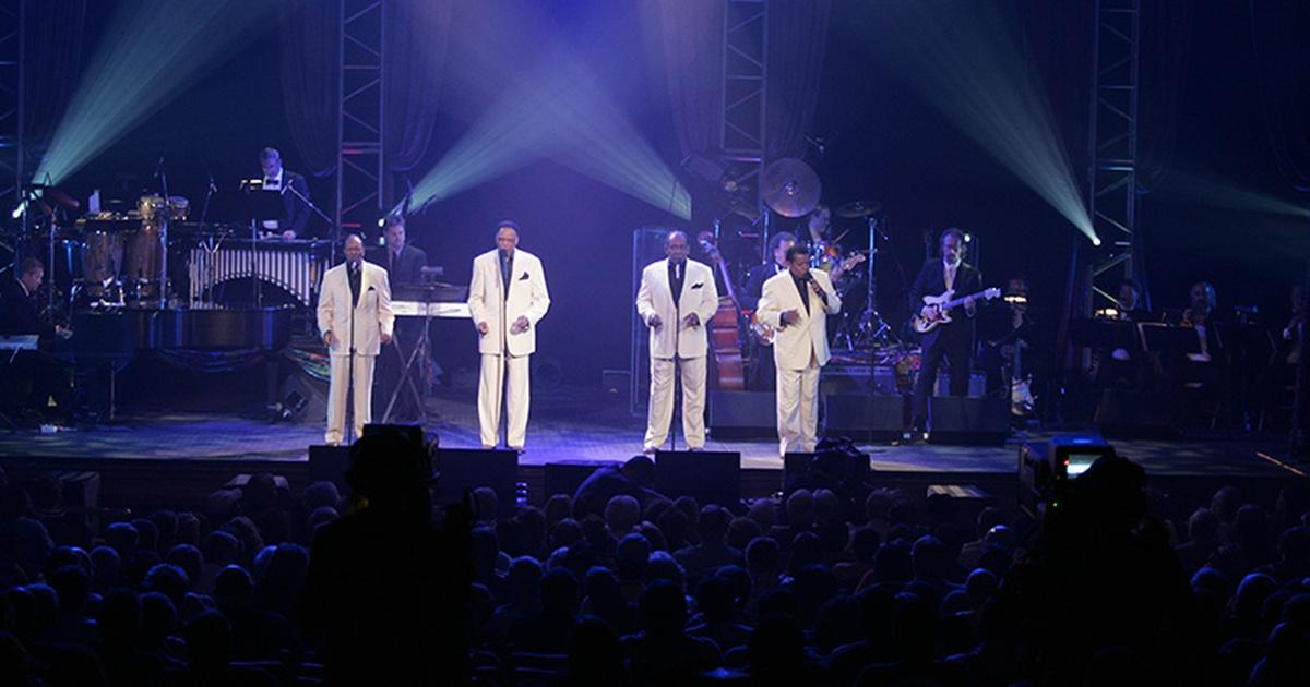 Four Black musicians wearing white suits and dark button-down shirts sing on stage.