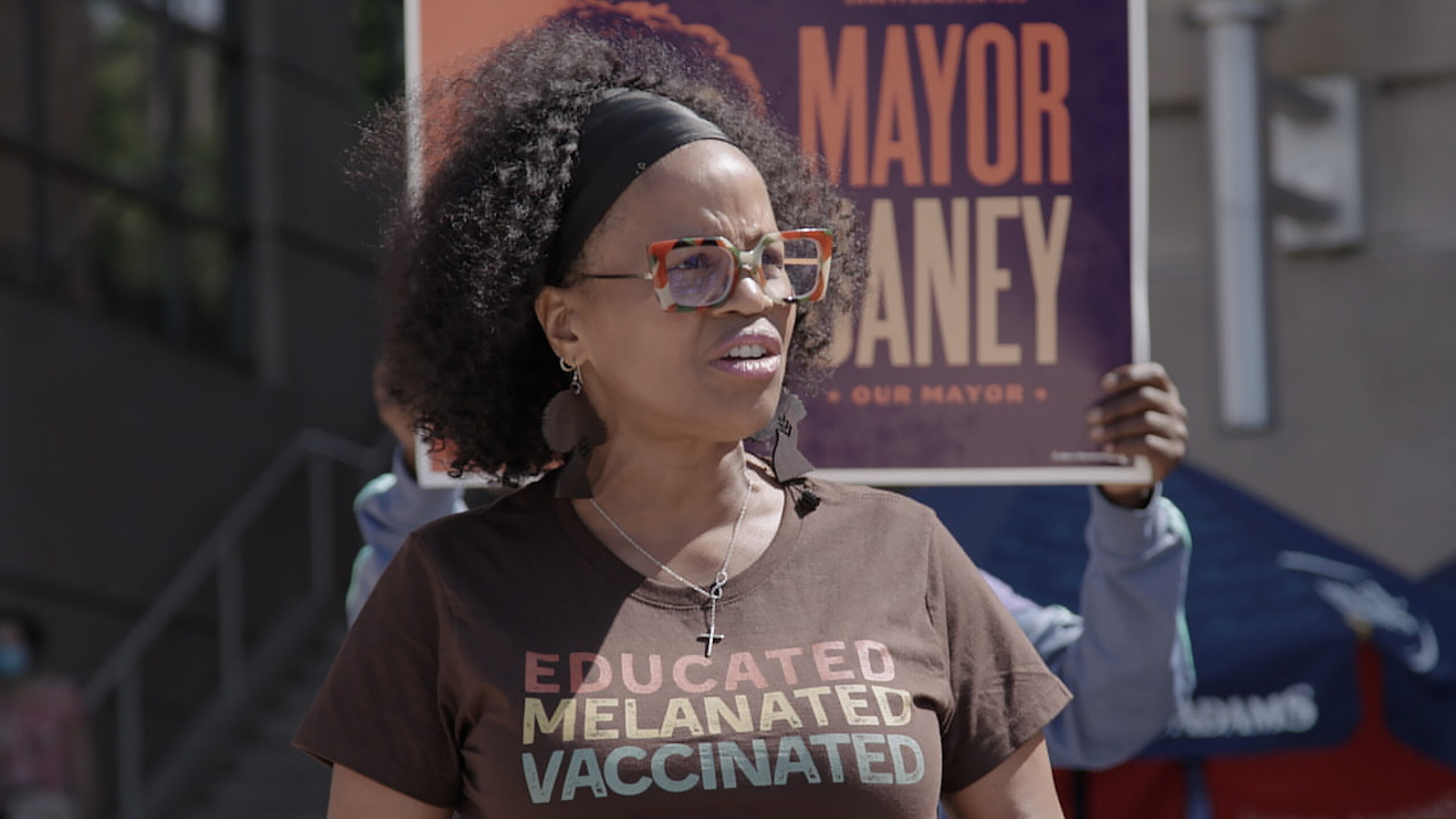 A Black woman with short Black hair pulled back with a dark scarf wears a brown t-shirt with type on the front of it.