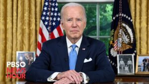 President Biden, an older white male with short gray hair wearing a blue suit, white shirt and light blue tie sits at a desk with his hands folded. The American flag stands in the background.