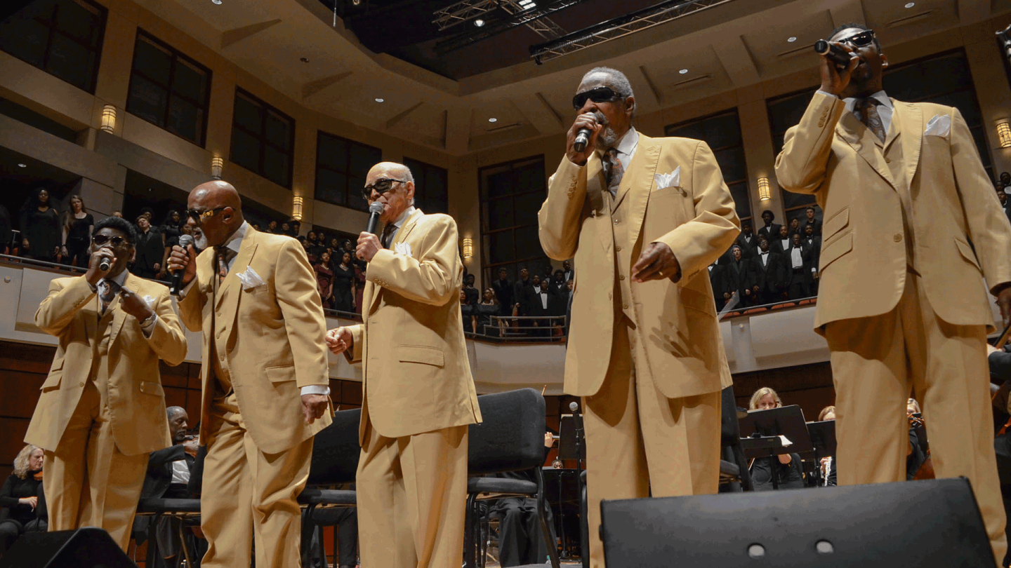 5 old men singing on stage in matching suites