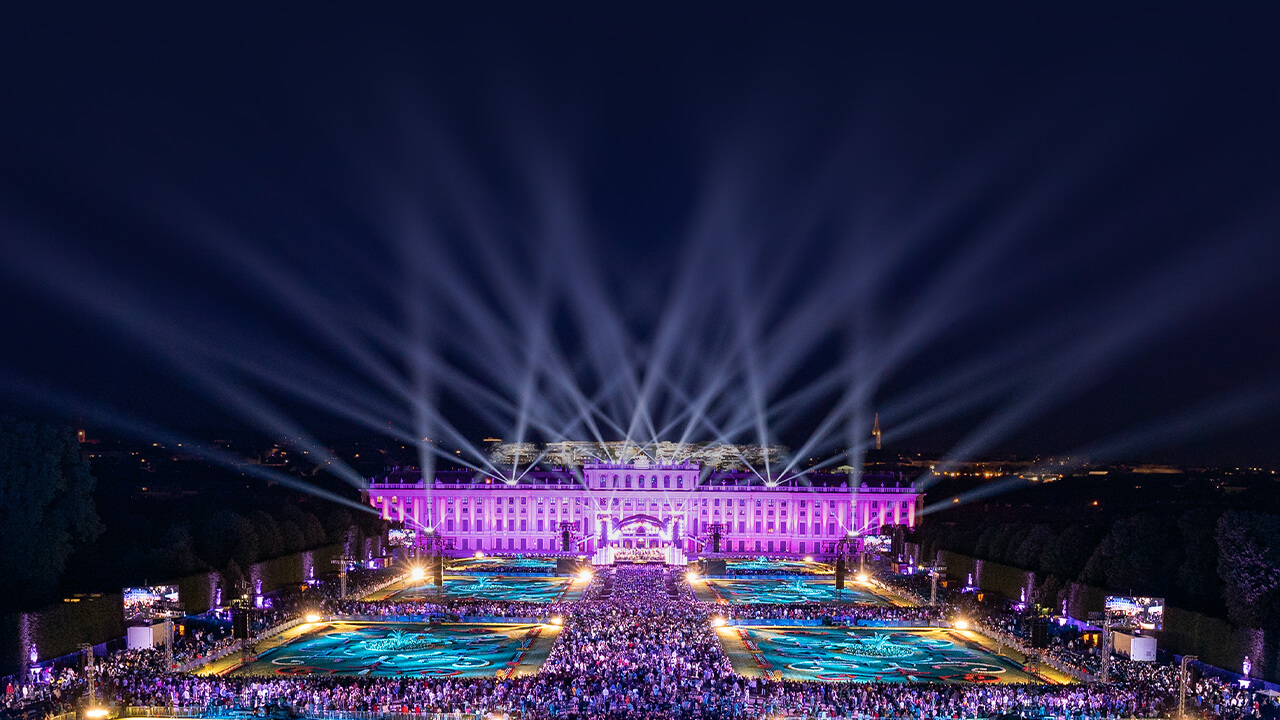 Vienna’s Schönbrunn Palace Gardens - a large building lit up in a purple light with spotlights shooting up in the air of a starry sky