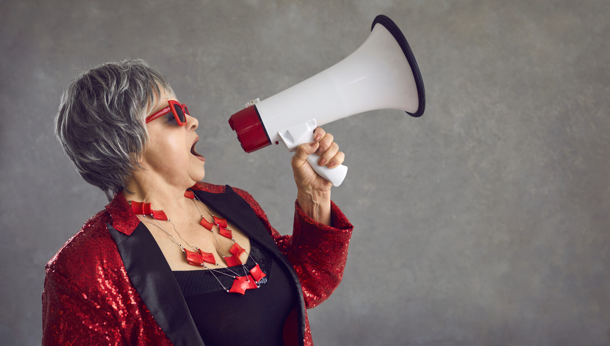 An older woman with short gray hair wearing a Black and red dress and red frames sun glasses yells into a megaphone.