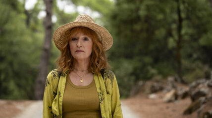 Lesley Manville stands outside wearing a straw hat and green blouse with a brown t-shirt underneath