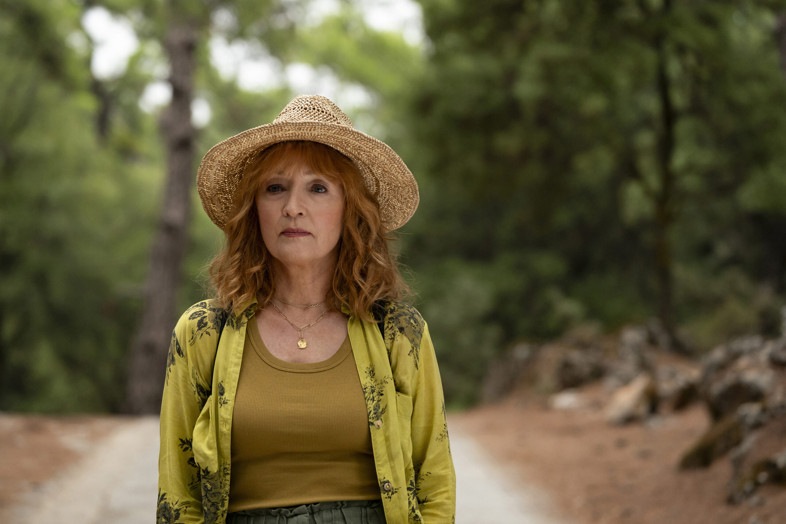 Lesley Manville stands outside wearing a straw hat and green blouse with a brown t-shirt underneath