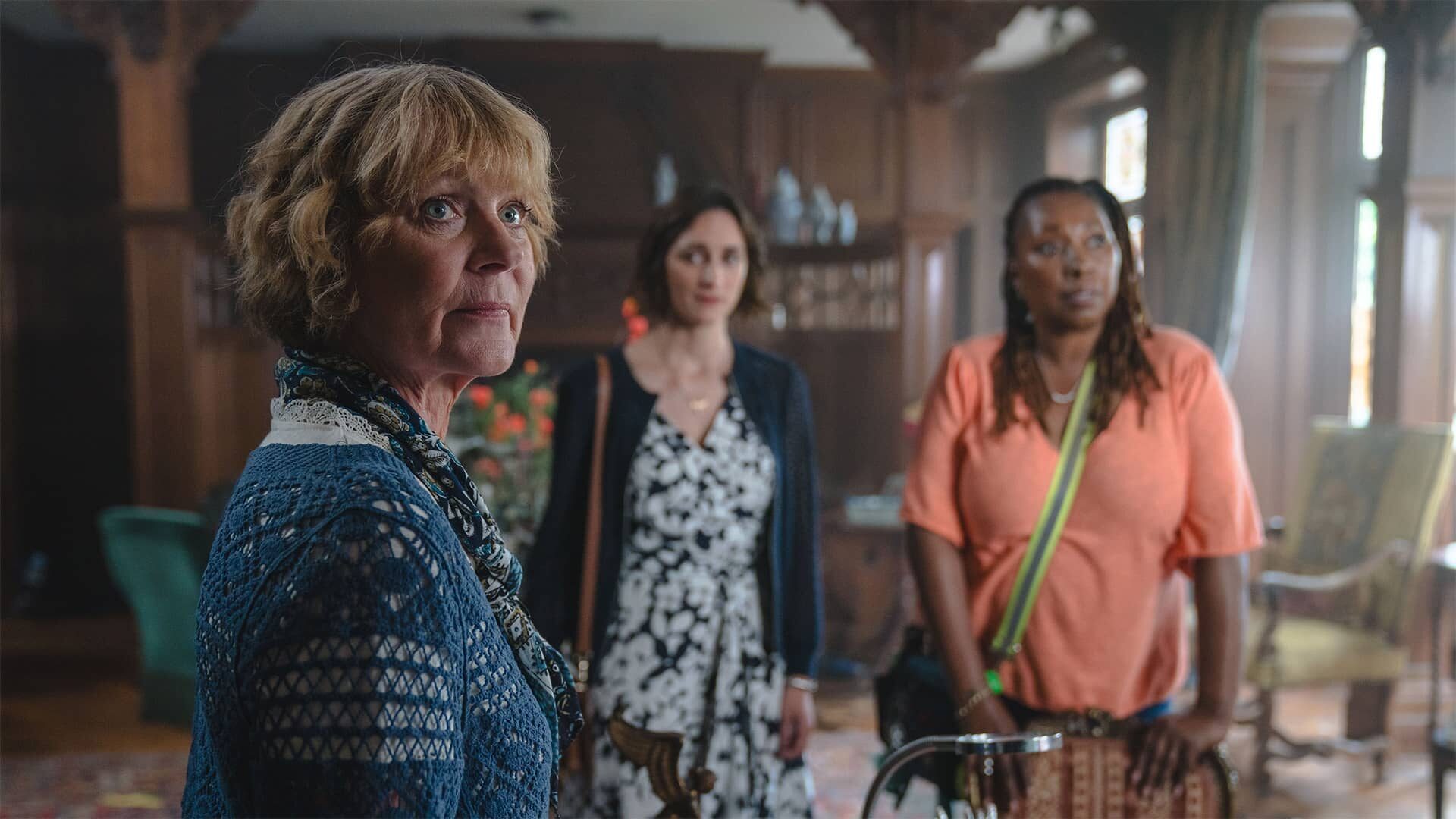 An older white woman with short blonde hair stands in the forefront while a white woman with short brown hair and a Black woman with long Black hair stands in the foreground.