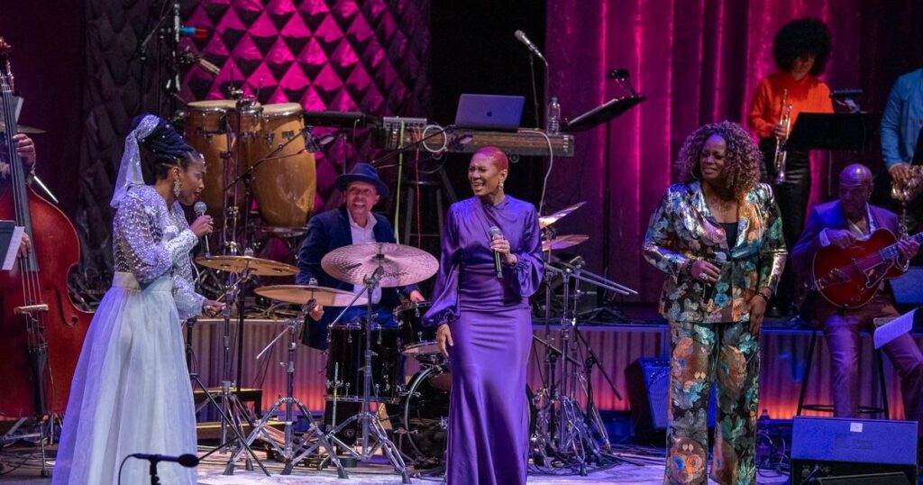 Three Black female singers known as the Baylor Project perform on stage with a fll band behind them.