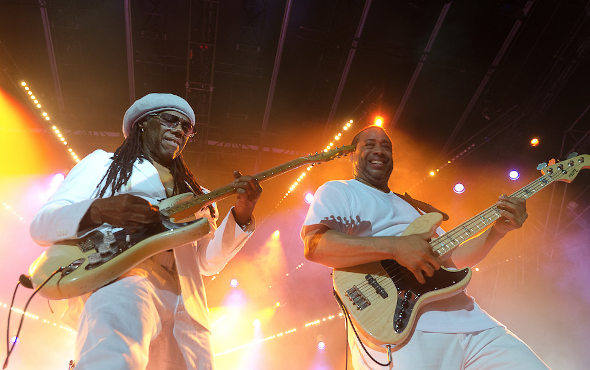 Two Black male musicians wearing white clothing stand on stage playing electric guitars.