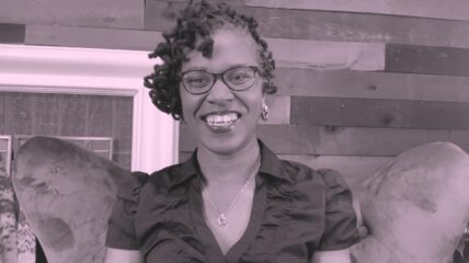 A black woman with short hair and glasses smiles into the camera