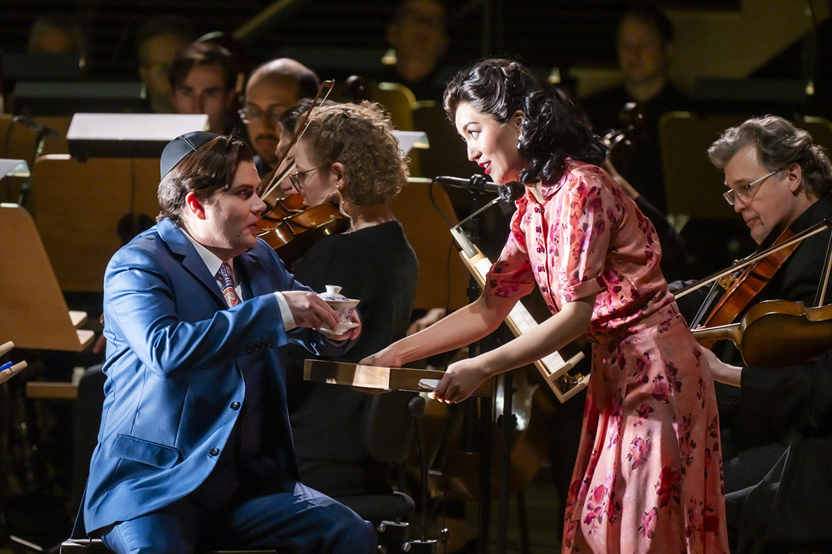 A young man in a blue suit wearing a yamaka takes a cup of tea handed to him by a woman with Black shoulder length hair wearing a red and shite print dress.