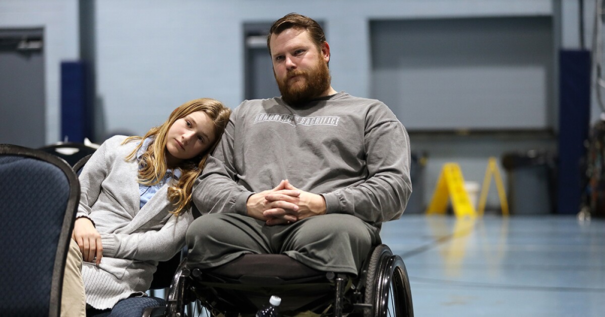 A white male in a gray sweatshirt sits in a wheel chair. His tween daughter sits beside him with her head on his shoulder