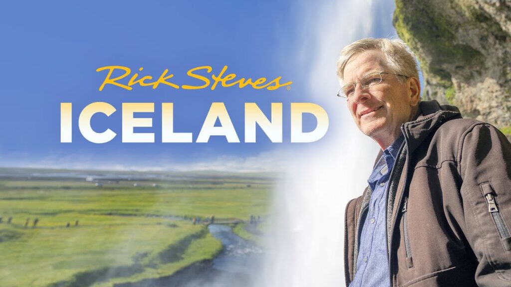 Rick Steves wearing a brown coat and blue button down stands in from of a blue sky, blue water and green grasses.