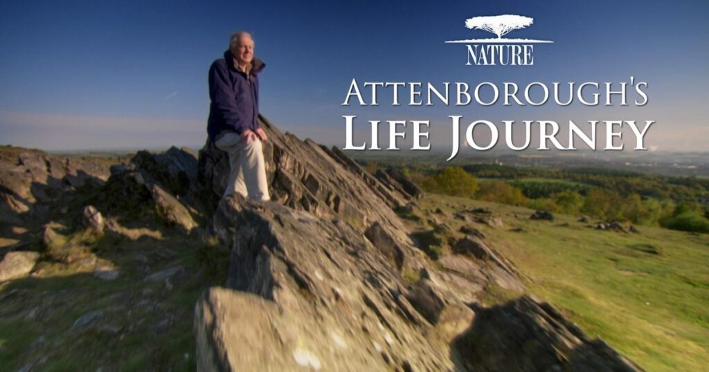 Sir David Attenborough wearing a blue jacket and white pants sits on top of a mountain.