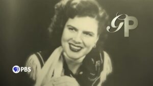 Patsy Cline wearing a white blouse and scarf with a dark embordered vest.