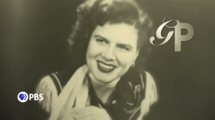 Patsy Cline wearing a white blouse and scarf with a dark embordered vest.