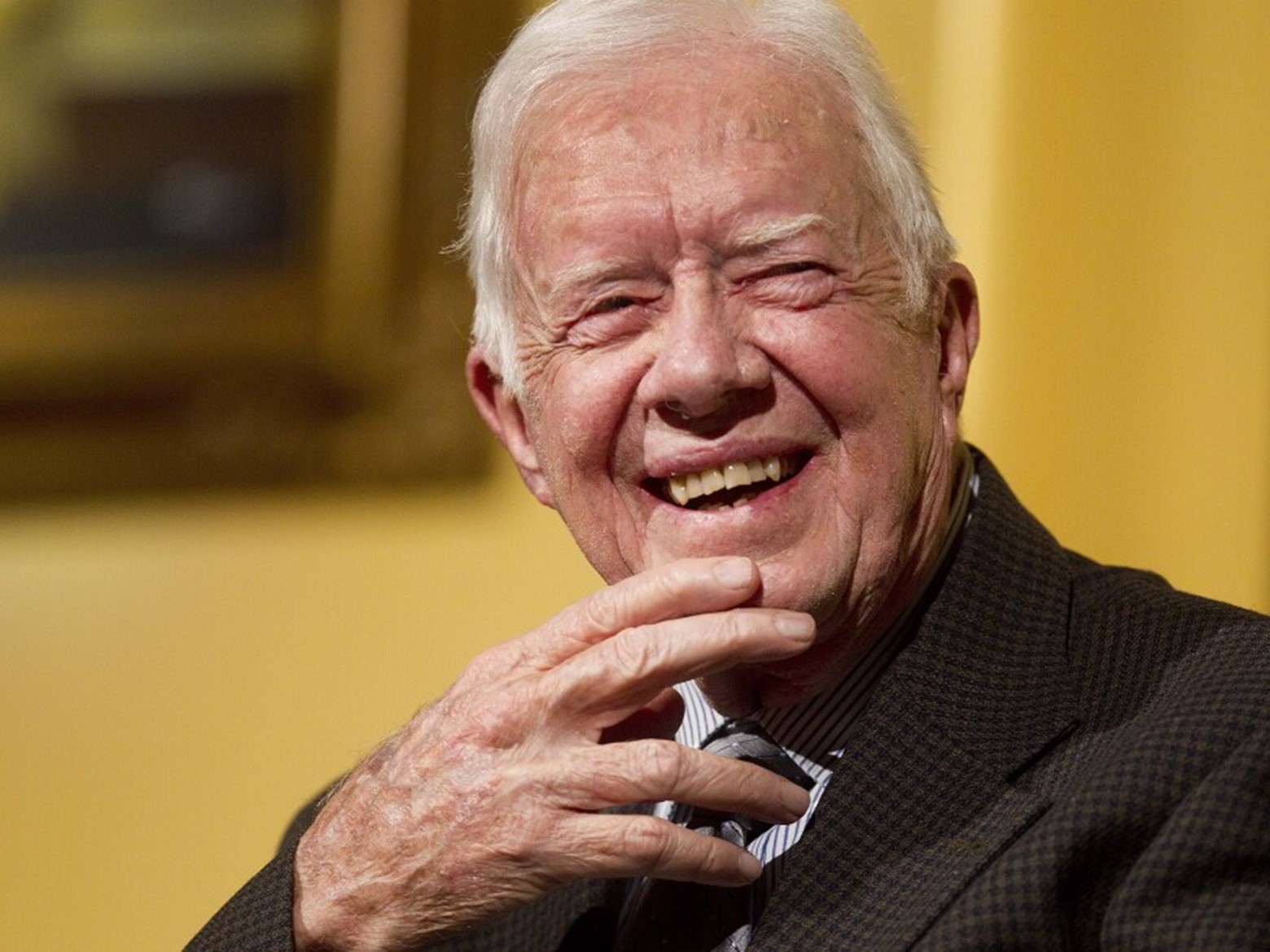 Former President Jimmy Carter in a Black and brown checkered blazer and gray shirt smiles with his hand on his chin.