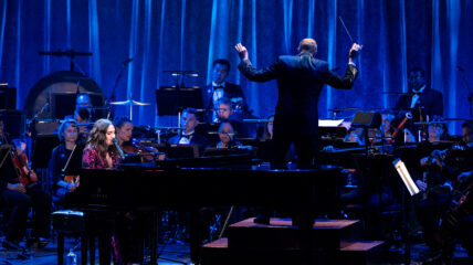 Singer Sara Bareilles wearing a Black and red dress sits at a piano and mic with an orchestra in the background