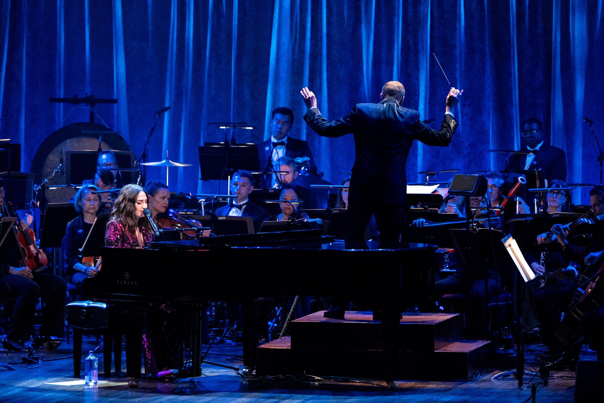 Singer Sara Bareilles wearing a Black and red dress sits at a piano and mic with an orchestra in the background