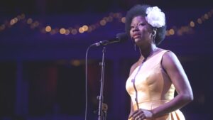 Singer Vanessa Hayes wearing a yellow dress and a white flower in her hair stands in front of a microphone.