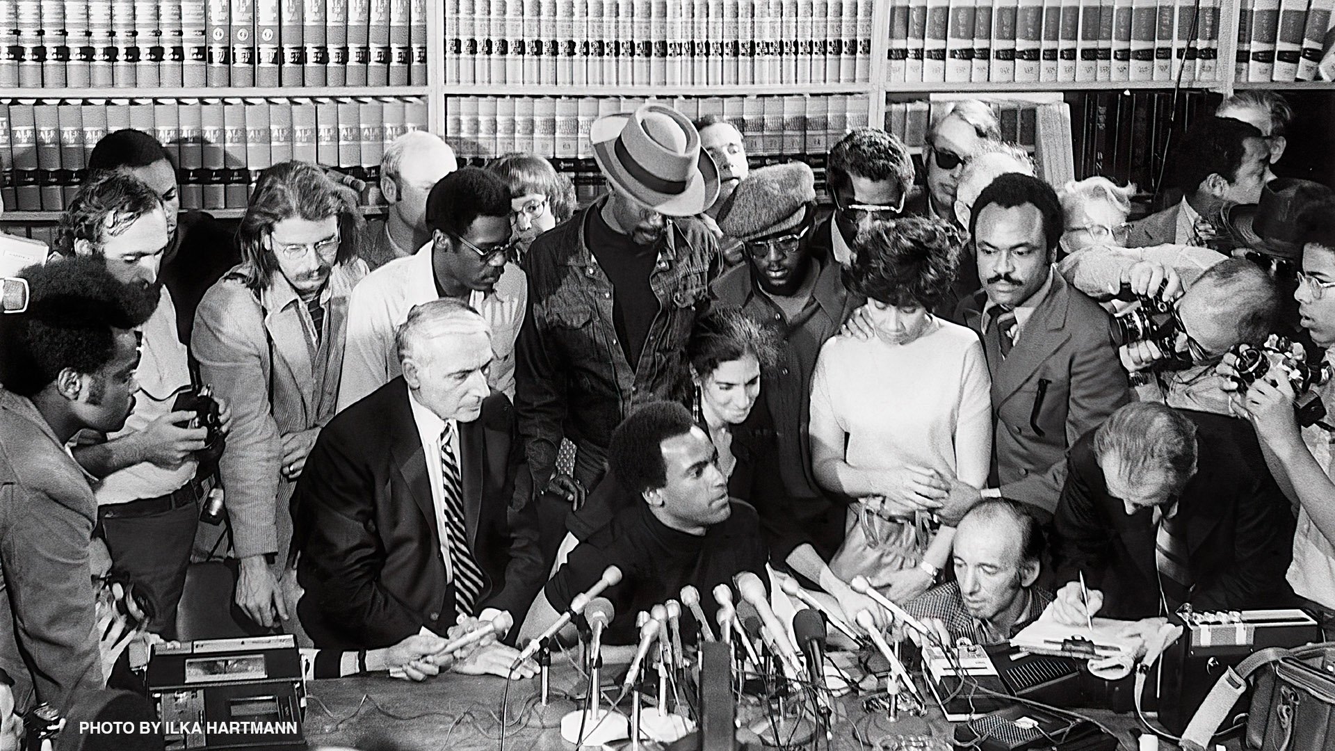 Huey Newton sits at a table at his press conference with people all around him,