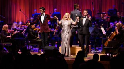 Michael Maliakel, Megan Hilty, and Luke Hawkins stand on a stage holding hands.