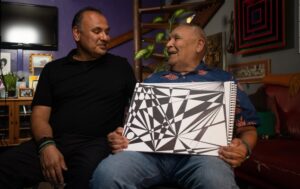 A grown son sits next to his father. The father is holding a drawing he has done.